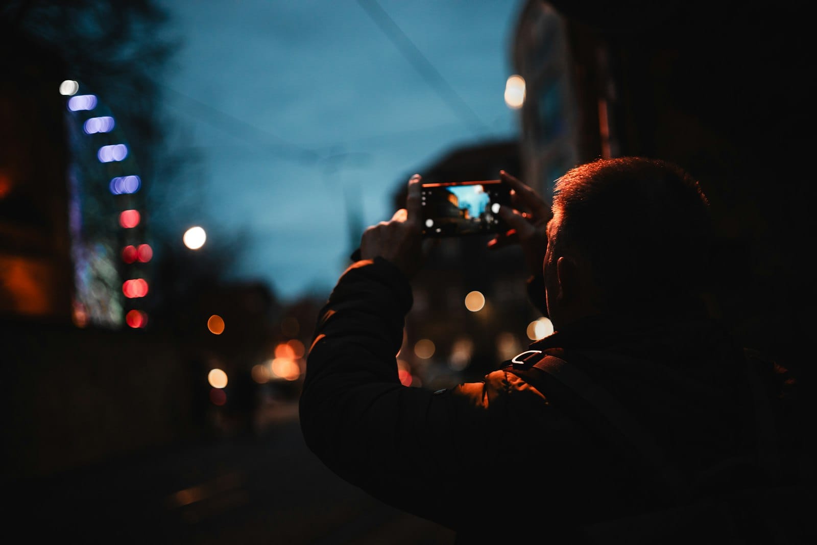 A person filming a city street at night on a smartphone, city lights blurred behind.