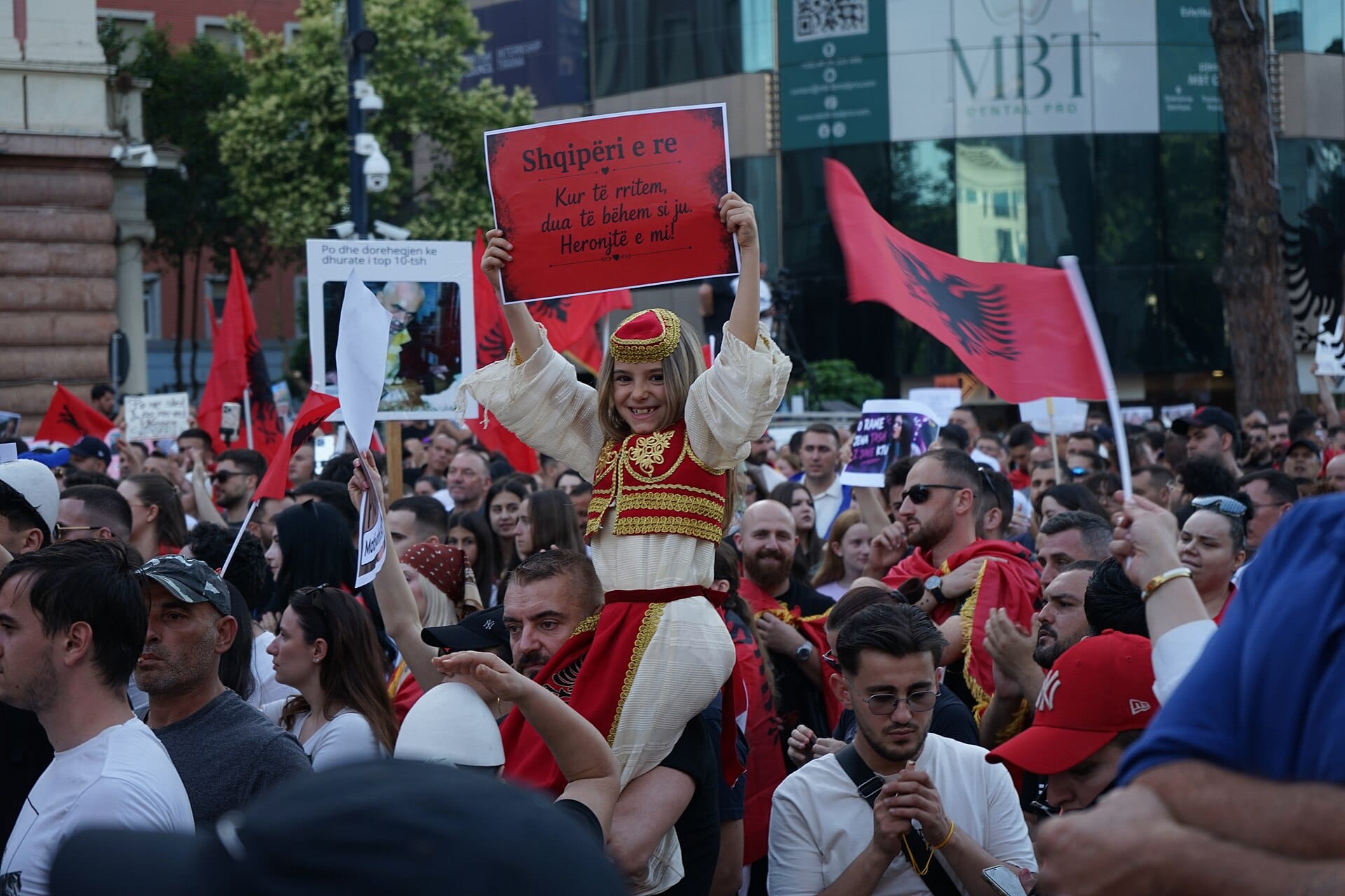 A girl in traditional Albanian dress holds up a sign at a Tirana rally, surrounded by people waving Albanian flags.