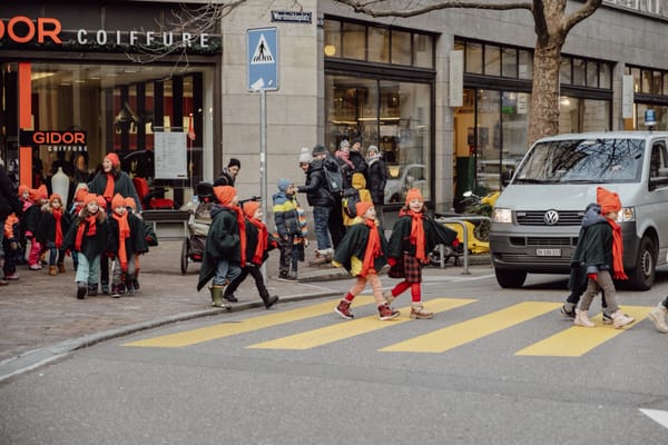 Eine Kindergartengruppe mit orangen Mützen, orangen Schals und dunkelgrünen Umhängen überquert in Zürich einen gelben Fussgängerstreifen